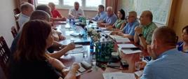 W sali konferencyjnej odbywa się spotkanie kilkunastu osób siedzących wokół długiego, drewnianego stołu. Uczestnicy dyskutują nad dokumentami rozłożonymi przed nimi. Na stole znajdują się kartki, notatniki, długopisy, telefony komórkowe, szklanki z wodą oraz kilka butelek wody mineralnej ustawionych na okrągłych tacach. W centralnej części stołu ustawiono niewielką kompozycję z białych kwiatów.
Na końcu stołu siedzi mężczyzna w białej koszuli, który sprawia wrażenie prowadzącego spotkanie. Pozostali uczestnicy zwracają uwagę w jego stronę lub prowadzą rozmowę z osobami siedzącymi naprzeciwko. Po prawej stronie stołu siedzą między innymi kobieta w zielonej bluzce oraz mężczyzna w zielonej koszuli w kratę, który wydaje się zabierać głos. W pomieszczeniu znajduje się około piętnastu osób – kobiet i mężczyzn.
W tle widoczne są duże okna z roletami przepuszczającymi światło dzienne, zasłony oraz duża mapa zawieszona na ścianie. Zdjęcie przedstawia formalne posiedzenie lub naradę w urzędzie albo instytucji, podczas której omawiane są dokumenty i prowadzone są dyskusje.