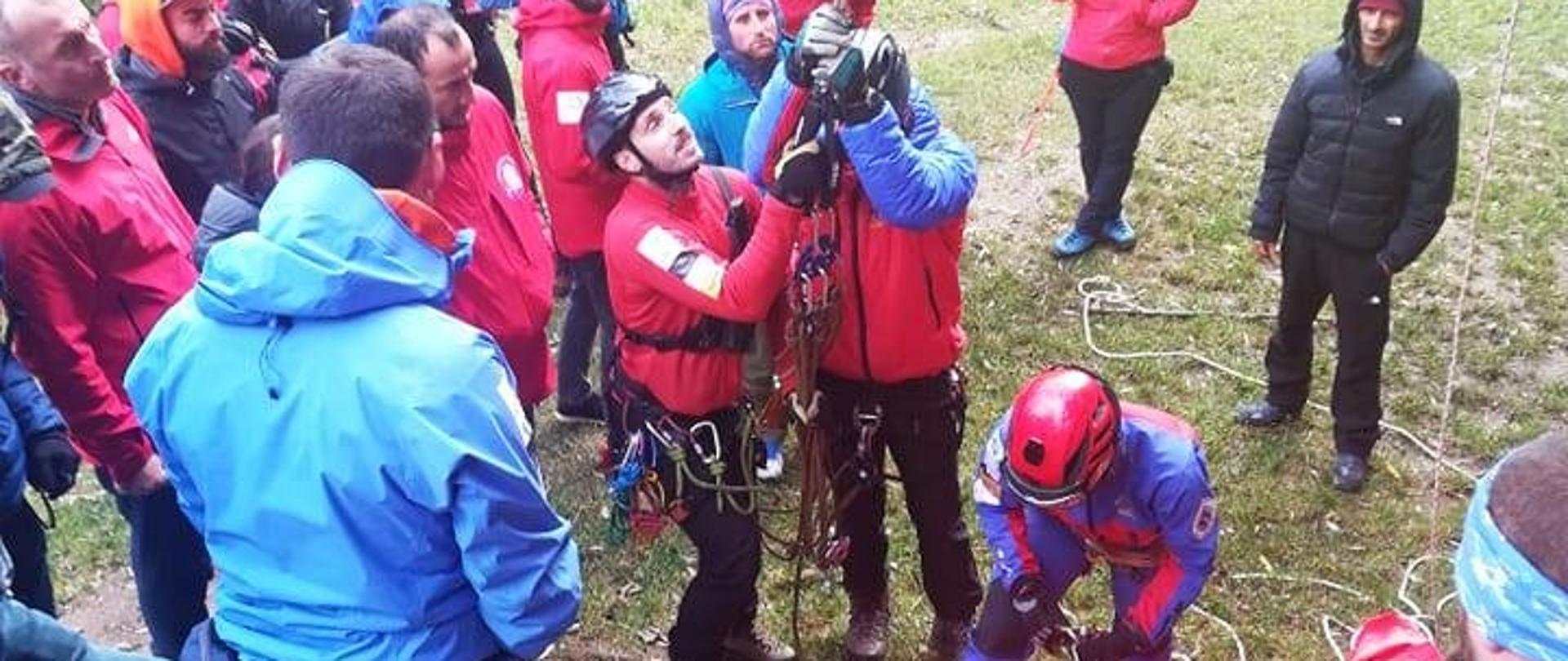 Grupa osób ubranych w czerwone i niebieskie kurtki trzyma linę.. jedna osoba leży na noszach udając osobę poszkodowaną w celu przeprowadzenia szkolenia ratowników górskich
