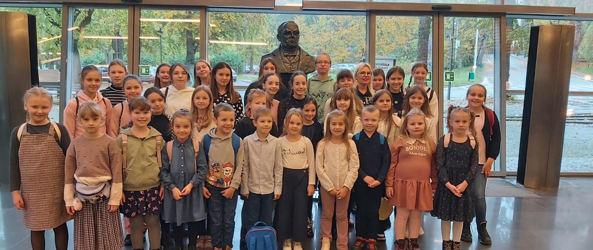 Uczniowie stoją w holu Filharmonii Koszalińskiej na tle pomniku Stanisława Moniuszki