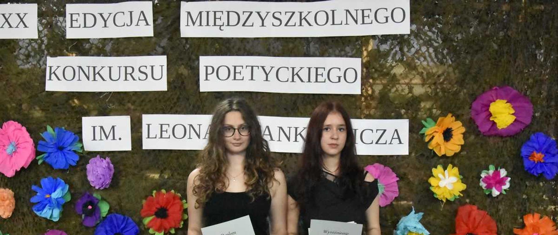 Zdjęcie przedstawia dwie młode kobiety stojące obok siebie na tle dużej, ozdobnej dekoracji ściennej. Obie uczestniczki patrzą prosto w stronę aparatu i trzymają w dłoniach otrzymane dokumenty.Po lewej stronie stoi dziewczyna o długich, kręconych, ciemnobrązowych włosach, nosząca okulary w ciemnych oprawkach. Jest ubrana w czarną bluzkę na ramiączkach i czarne spodnie. Oburącz trzyma przed sobą pionowo białą kartkę z napisem „Dyplom”.Po prawej stronie stoi druga dziewczyna z długimi, prostymi, ciemnymi włosami z przedziałkiem na środku. Ma na sobie czarną bluzkę z krótkim rękawem oraz czarne spodnie. W dłoniach trzyma białą kartkę z nagłówkiem „Wyróżnienie”.W tle znajduje się ciemnozielona siatka maskująca przypominająca mchy lub liście. Jest ona gęsto udekorowana dużymi, przestrzennymi kwiatami z kolorowej bibuły (w kolorach różowym, niebieskim, żółtym, pomarańczowym, fioletowym i czerwonym).Nad głowami dziewcząt, na dekoracji, rozmieszczone są prostokątne, białe paski papieru z czarnym, drukowanym napisem: „XX EDYCJA MIĘDZYSZKOLNEGO KONKURSU POETYCKIEGO IM. LEONA STANKIEWICZA”.W dolnych rogach kadru widoczne są elementy dodatkowych dekoracji: po lewej stronie biała rączka wiklinowego koszyka, a po prawej stronie fragmenty drewnianych konstrukcji z brzozowych pieńków ozdobionych sztucznymi słonecznikami.