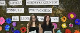 Zdjęcie przedstawia dwie młode kobiety stojące obok siebie na tle dużej, ozdobnej dekoracji ściennej. Obie uczestniczki patrzą prosto w stronę aparatu i trzymają w dłoniach otrzymane dokumenty.Po lewej stronie stoi dziewczyna o długich, kręconych, ciemnobrązowych włosach, nosząca okulary w ciemnych oprawkach. Jest ubrana w czarną bluzkę na ramiączkach i czarne spodnie. Oburącz trzyma przed sobą pionowo białą kartkę z napisem „Dyplom”.Po prawej stronie stoi druga dziewczyna z długimi, prostymi, ciemnymi włosami z przedziałkiem na środku. Ma na sobie czarną bluzkę z krótkim rękawem oraz czarne spodnie. W dłoniach trzyma białą kartkę z nagłówkiem „Wyróżnienie”.W tle znajduje się ciemnozielona siatka maskująca przypominająca mchy lub liście. Jest ona gęsto udekorowana dużymi, przestrzennymi kwiatami z kolorowej bibuły (w kolorach różowym, niebieskim, żółtym, pomarańczowym, fioletowym i czerwonym).Nad głowami dziewcząt, na dekoracji, rozmieszczone są prostokątne, białe paski papieru z czarnym, drukowanym napisem: „XX EDYCJA MIĘDZYSZKOLNEGO KONKURSU POETYCKIEGO IM. LEONA STANKIEWICZA”.W dolnych rogach kadru widoczne są elementy dodatkowych dekoracji: po lewej stronie biała rączka wiklinowego koszyka, a po prawej stronie fragmenty drewnianych konstrukcji z brzozowych pieńków ozdobionych sztucznymi słonecznikami.