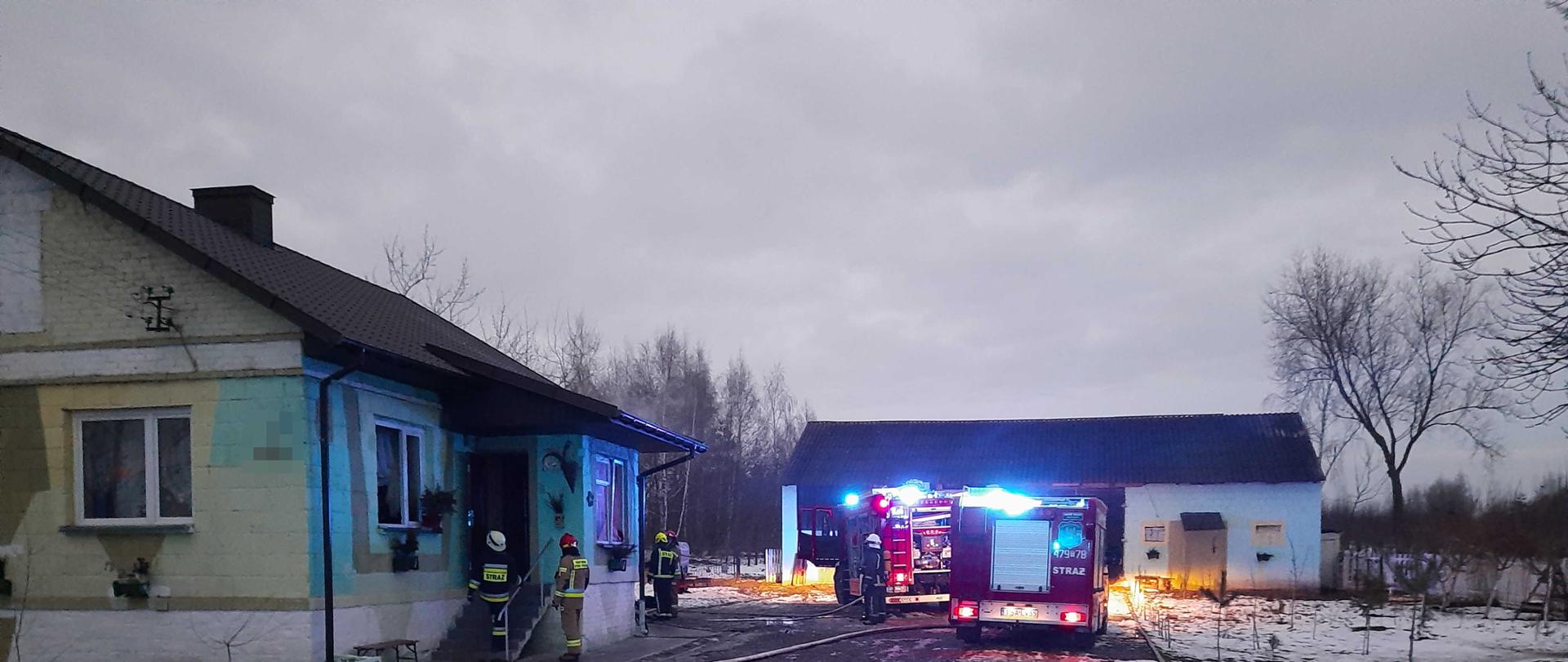 Zdjęcie zrobione na tle budynku mieszkalnego i gospodarczego z ośnieżonym podjazdem. Na zdjęciu widać dwa samochody strażackie i rozwinięty wąż strażacki. Obok samochodów strażackich widać strażaków, którzy gaszą pomieszczenie kotłowni i sprawdzają budynek mieszkalny. 