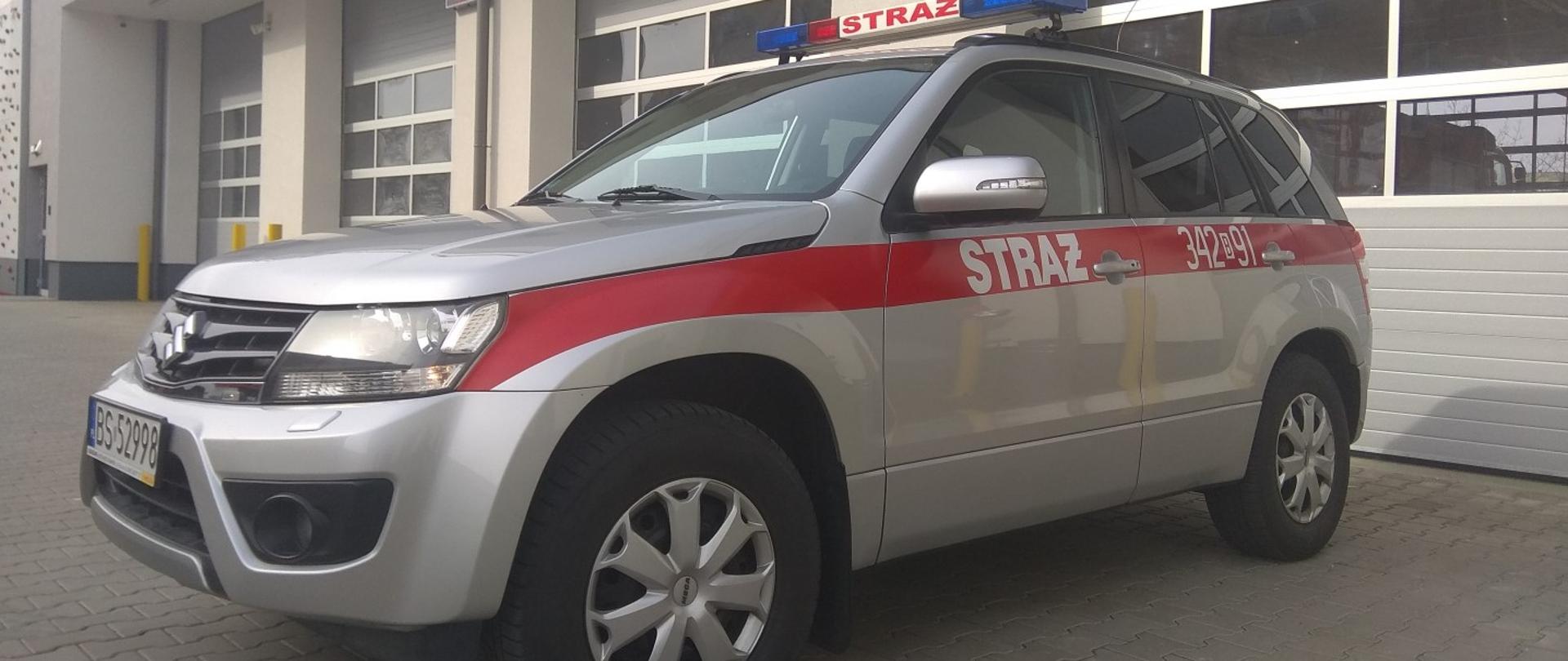 Na pierwszym planie samochód osobowy koloru szarego z czerwonym paskiem pośrodku pojazdu. W tle zdjęcia garaże. 