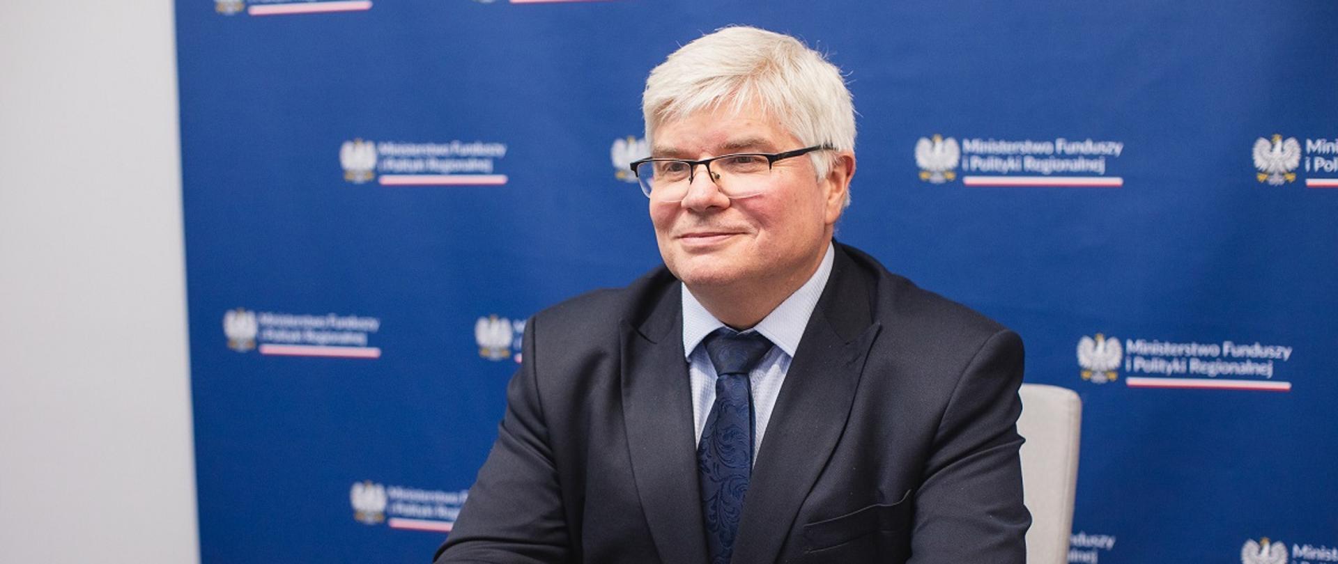 Wiceminister funduszy i polityki regionalnej Maciej Lasek siedzi przy stole, za jego plecami ścianka z logotypem MFiPR