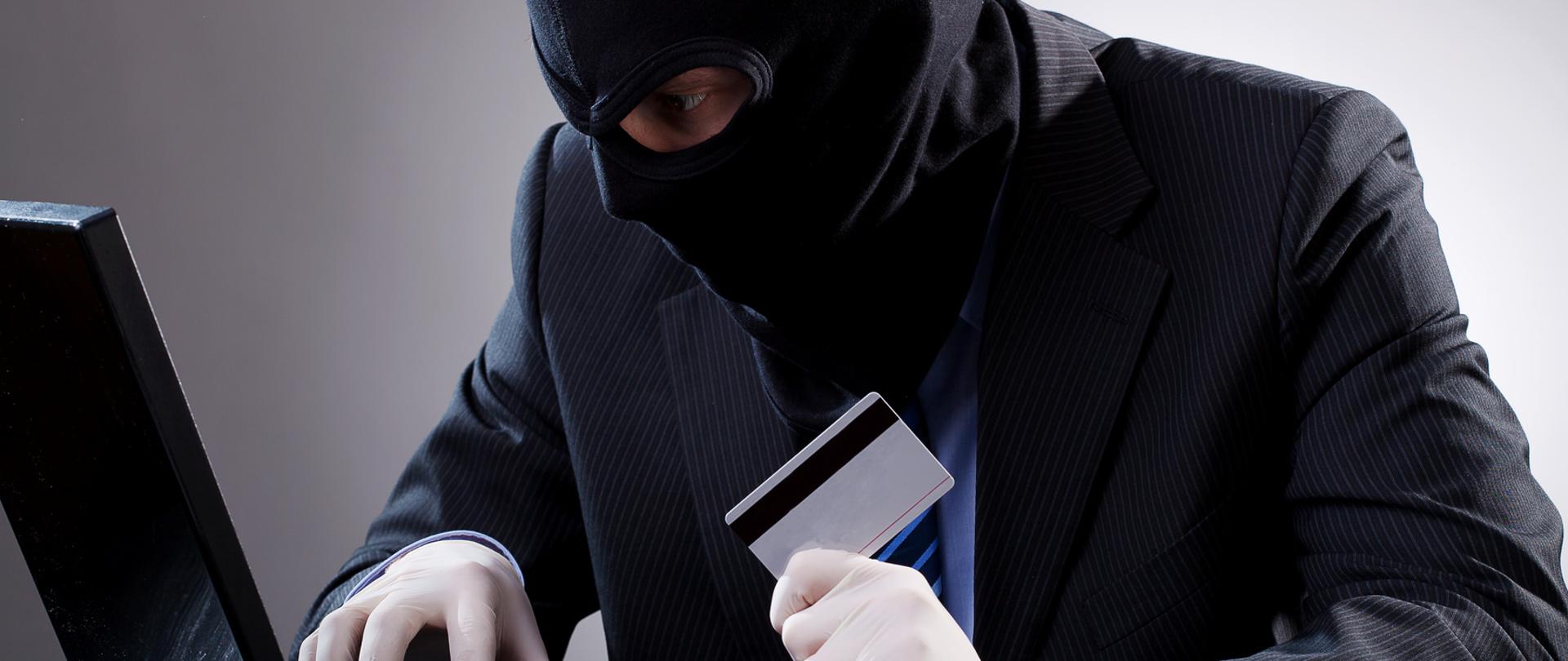 Mężczyzna w kominiarce i rękawiczkach z kartą kredytową przy komputerze.