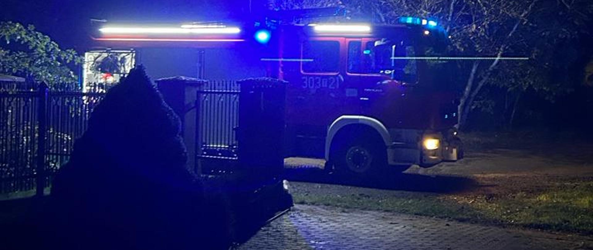 Zdjęcie przedstawia samochód pożarniczy stojący przy otwartej bramie wjazdowej na posesję gdzie doszło do pożaru.
