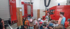 Zdjęcie przedstawia grupę dzieci podczas zapoznania ze sprzętem strażackim w pomieszczeniu garażu na tle pojazdów strażackich koloru czerwonego.