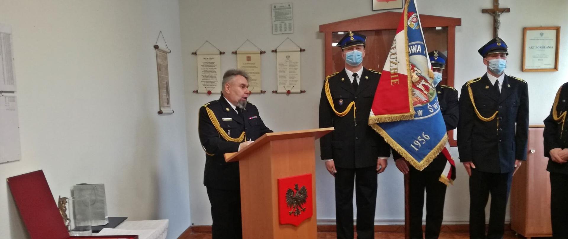 Zdjęcie przedstawia 5 mężczyzn ubranych w czarne garnitury z ozdobnym złotym sznurem. Obok niego znajduję się stół z prezentami, poczet sztandarowy KP PSP Świdwin, mównica przed którą przemawia odchodzący na emeryturę strażak, Godło Rzeczypospolitej Polskiej oraz Krzyż.