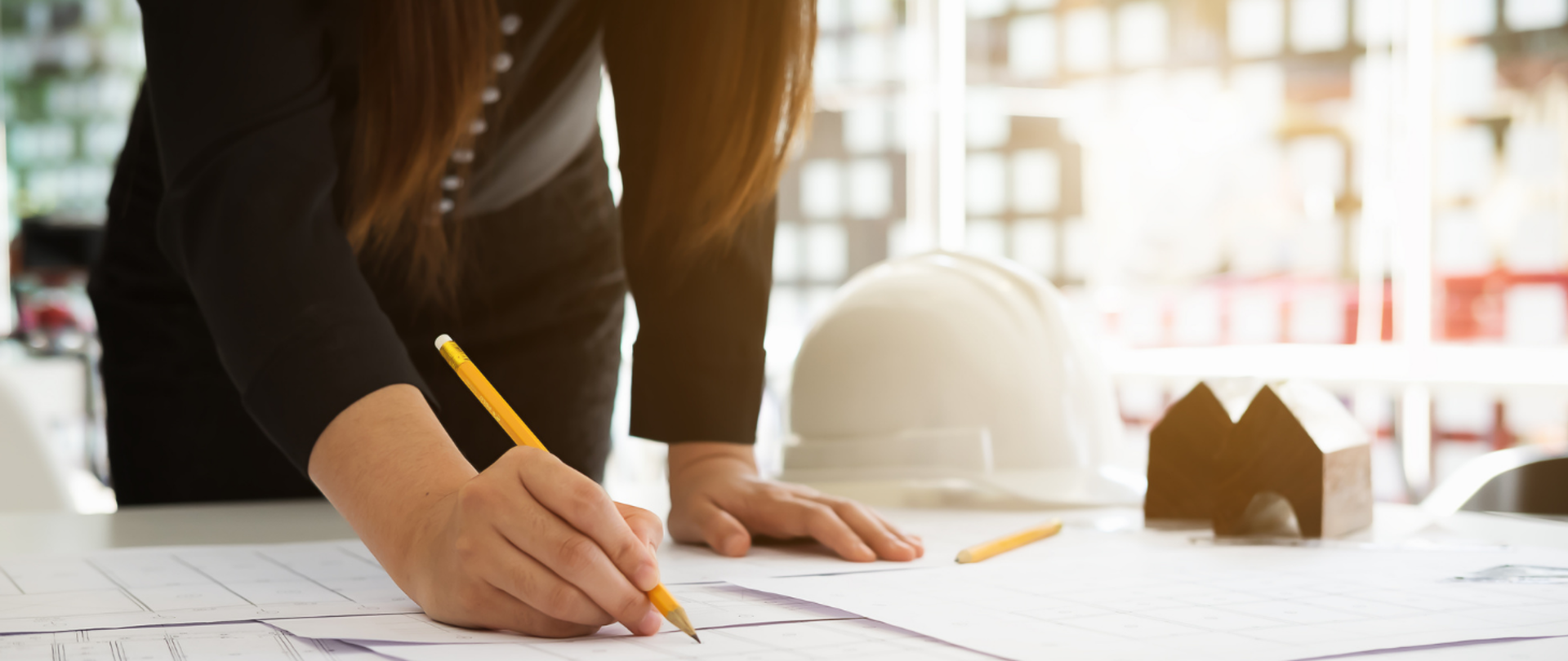 Kobieta pisze ołówkiem po zadrukowanych kartkach papieru leżących na stole. Obok jej lewej dłoni leży biały kask, żółty ołówek oraz makieta urbanistyczna.