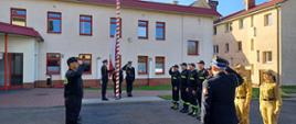 Uroczystość z okazji Dnia Flagi Rzeczypospolitej Polskiej. Strażacy ze zmian służbowych i funkcjonariusze z komendy stoją naprzeciwko siebie. Komendant, dowódcy zmian i asystujący pocztu flagowego salutują.