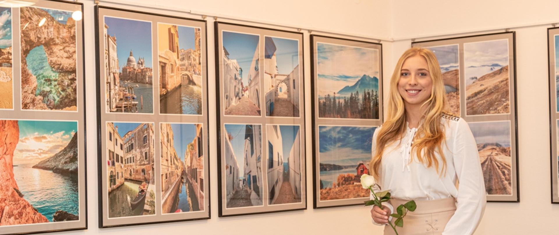 Na zdjęciu autorka wystawy fotograficznej Klaudia Glazar stojąca na tle swoich zdjęć, które przedstawiają krajobrazy. Autorka w rękach trzyma białą różę. 