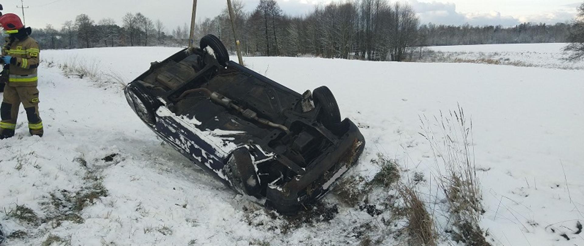 Na zdjęciu widoczne jest przewrócone na dach auto osobowe koloru granatowego. Pojazd znajduje się na polu pokrytym śniegiem w centralnej części zdjęcia. Przód pojazdu skierowany do oglądającego. Z lewej strony zdjęcia stoi strażak w ubraniu specjalnym koloru żółtego z czerwonym hełmem na głowie.