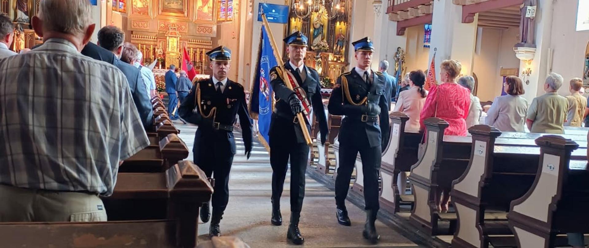 Zdjęcie przedstawia wnętrze kościoła, w którym główną alejką idzie poczet sztandarowy złożony z trzech mundurowych. Mężczyźni w czarnych garniturach galowych, czapkach z daszkiem i białych koszulach maszerują w równym szyku. Środkowy z nich niesie oparty na ramieniu sztandar z błękitnym płótnem, a pozostali dwaj asystują mu po bokach. Po obu stronach alejki, w drewnianych ławkach oraz w głębi świątyni, stoją zgromadzeni ludzie obserwujący uroczystość.