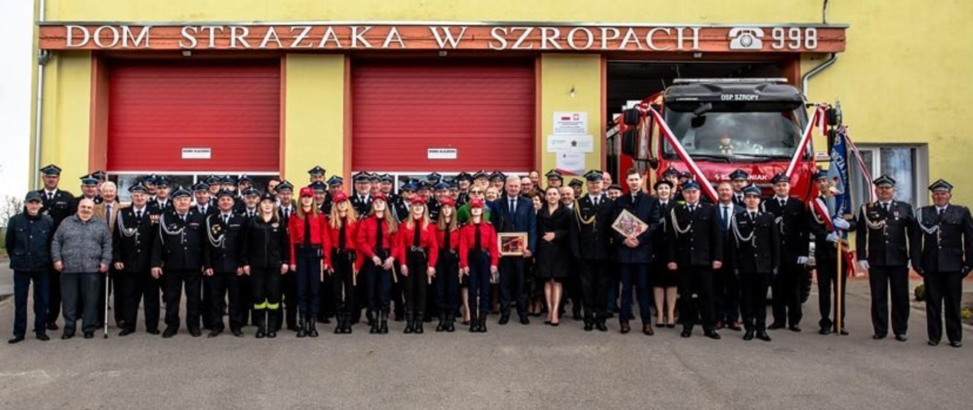 pora dzienna, przed budynkiem remizy Osp Szropy stoją tłumnie zgromadzenia goście, druhny i druhowie OSP Szropy. Po prawej stronie widać fragment przekazywanego pojazdu który na przedzie ustrojony jest szrafą w barwach narodowych.