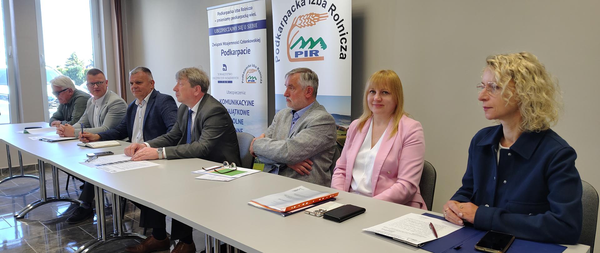 Pięć osób siedzi przy stole konferencyjnym podczas oficjalnego spotkania organizacyjnego. Po lewej stronie siedzi mężczyzna w ciemnej marynarce zapisujący notatki w notesie. Obok niego znajduje się mężczyzna w garniturze i krawacie, siedzący centralnie przed roll-upem z logo Podkarpackiej Izby Rolniczej. Dalej siedzi starszy mężczyzna w jasnej marynarce przeglądający dokumenty. Po prawej stronie widoczne są dwie kobiety – jedna ubrana w różowy garnitur, druga w granatowy komplet – obie siedzą skupione przy stole z dokumentami i teczkami. W tle znajdują się stojące banery informacyjne związane z Podkarpacką Izbą Rolniczą i ubezpieczeniami wzajemnymi. Sala jest nowoczesna, utrzymana w jasnych kolorach i dobrze oświetlona.