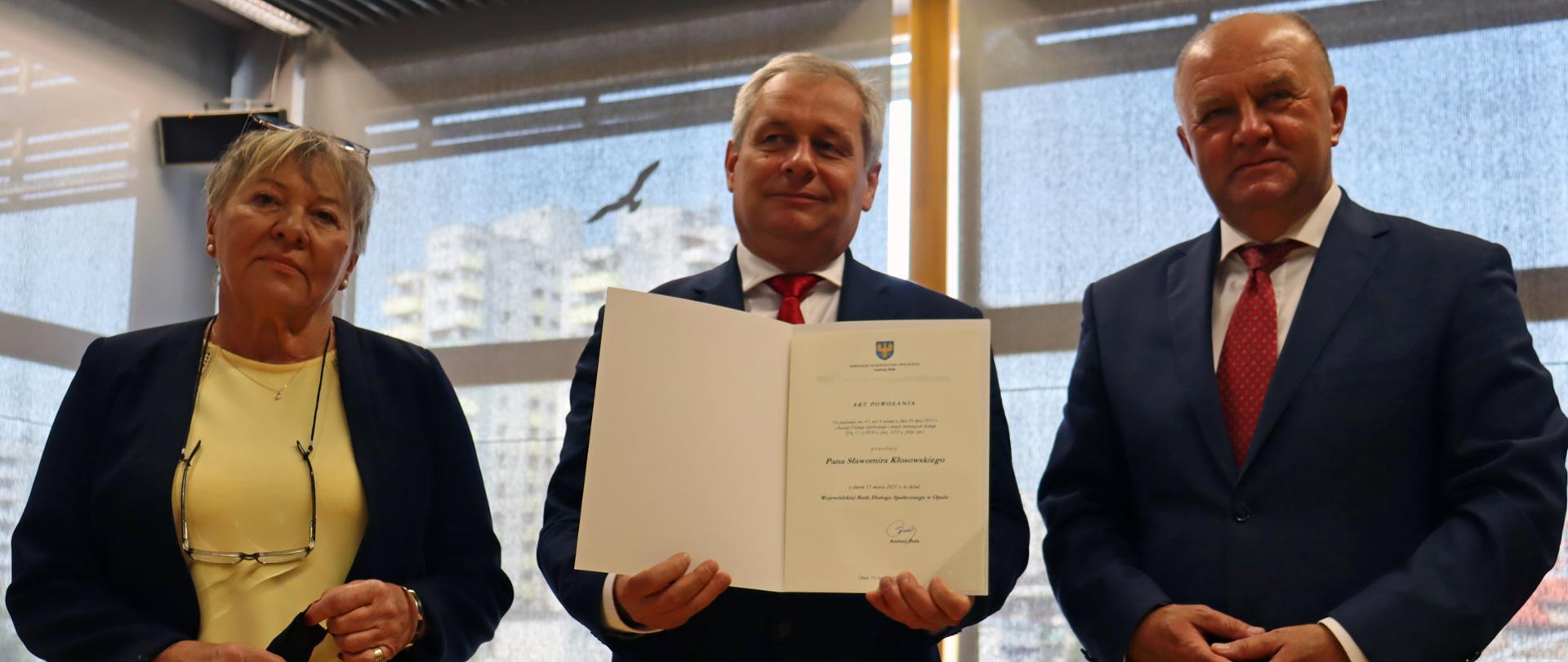 Wojewoda opolski Sławomir Kłosowski odebrał dziś akt powołania na członka opolskiej WRDS. Na zdjęciu trzy osoby. Od lewej przewodnicząca WRDS, ubrana w żółtą sukienkę i czarny żakiet. To starsza kobieta spoglądająca w bok kadru. Na jej szyi łańcuszek z dopiętymi okularami optycznymi, w ręku trzyma maseczkę medyczną. Obok niej - stojący w centrum wojewoda opolski, trzymający w dłoniach teczkę z aktem powołania. Po prawej stronie kadru, obok wojewody stoi marszałek regionu, który pozuje do zdjęcia, ma splecione dłonie W tle wystrój sali, w której odbyła się uroczystośc - widać duże, zasłonięte roletą okno oraz fragment biurka.
