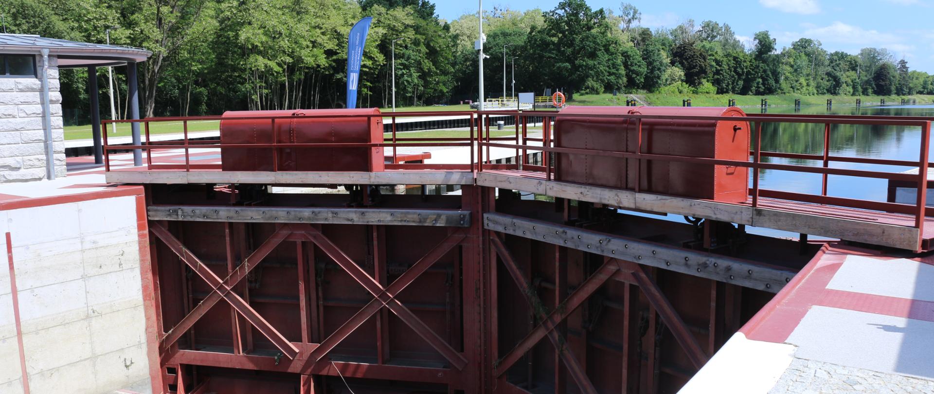Na zdjęciu śluza w m. Sławięcice w pow. kędzierzyńsko-kozielskim. Na pierwszym planie widać wielkie, brązowe i metalowe drzwi - to część śluzy. Powyżej nich widać taflę wody. W tle zieleń. 