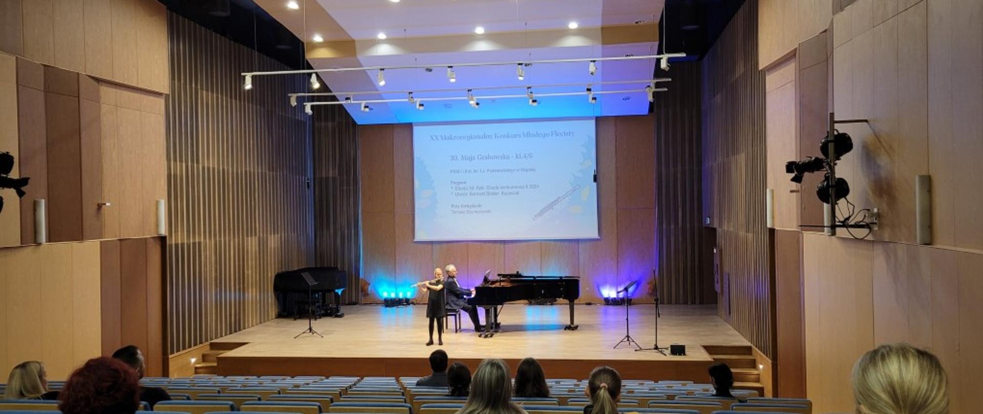 Zdjęcie kolorowe. Dziewczynka grająca na flecie poprzecznym w sali koncertowej szkoły muzycznej wraz z pianistą.