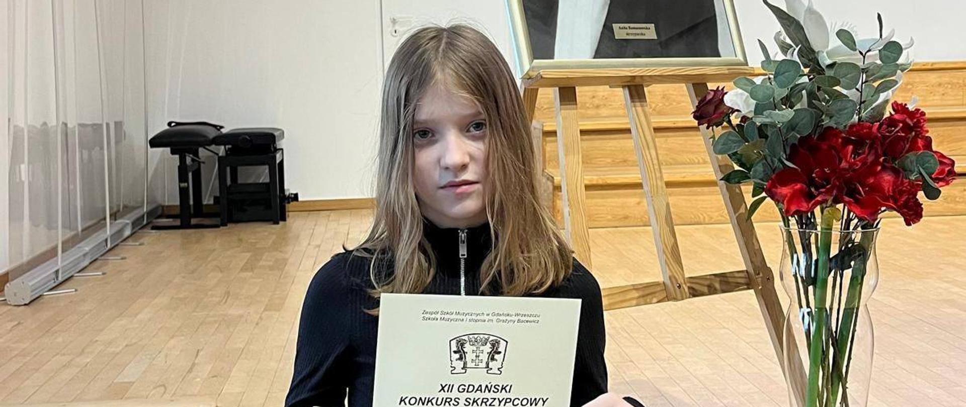 Portretowe ujęcie młodej dziewczynki stojącej na drewnianej scenie. Dziewczynka trzyma przed sobą dyplom z napisem „WYRÓŻNIENIE otrzymuje Julia Ostrowska”. W tle, tuż za nią, widoczna jest sztaluga z portretem Anity Romanowskiej oraz elegancki bukiet kwiatów.