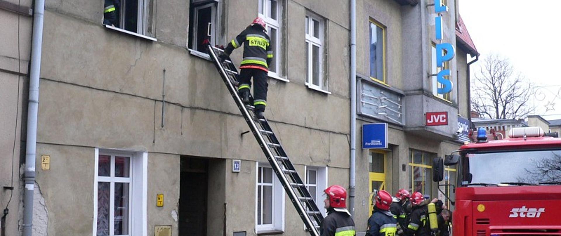 Widoczny budynek mieszkalny wielorodzinny dwu piętrowy. Przed budynkiem stoją strażacy przy samochodzie strażackim. Jeden strażak stoi na drabinie. Na ziemi widoczne nadpalone części wyposażenia mieszkania.