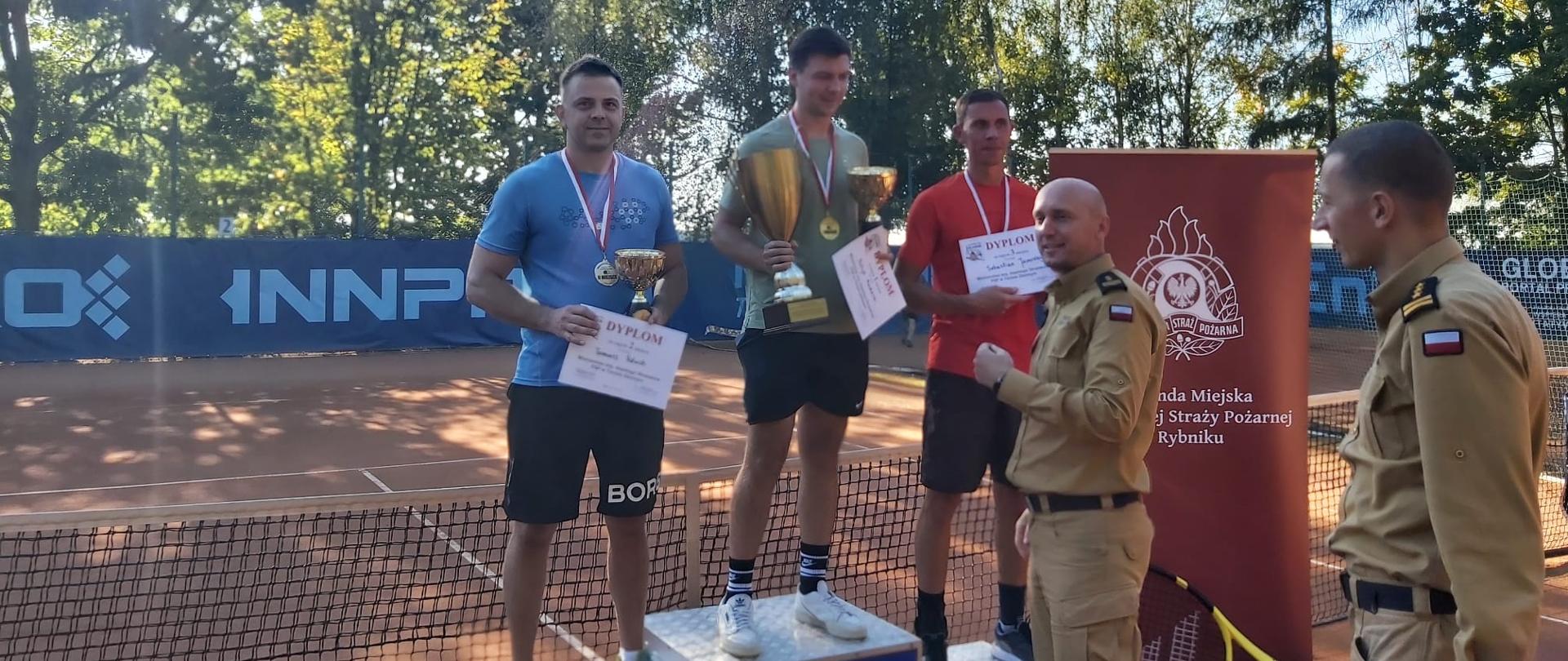 Zawodnicy z dyplomami w rękach i medalami na szyjach stoją na podium medalowym. Dwóch strażaków w mundurach stoi przed nimi. Obok nich stoi baner z logo PSP i napisem Komenda Miejska Państowej Straży Pożarnej w Rybniku. Za nimi niebieski baner na ogrodzeniu kortu i wysokie drzewa. 