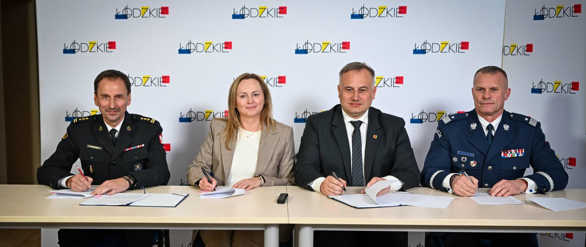 Czworo osób (policjant, kobieta w żakiecie, urzędnik i strażak) siedzi przy stole i jednocześnie podpisuje dokumenty, patrząc na kamerę. Tło stanowi baner z logo województwa łódzkiego.