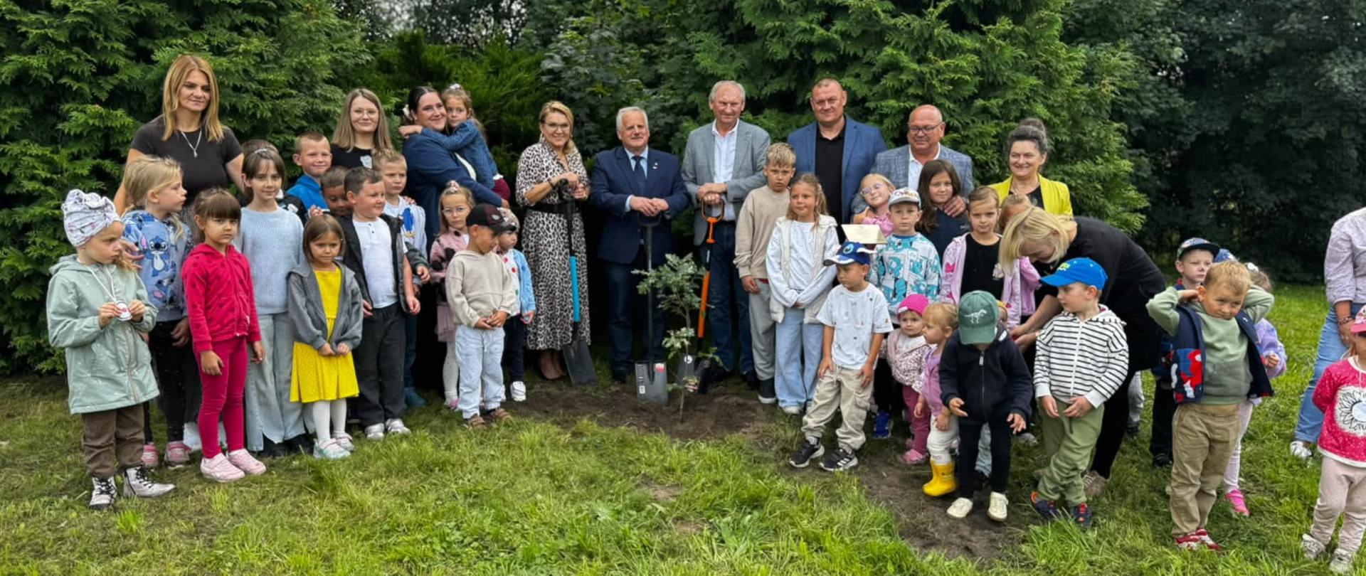 Grupa przedstawicieli władz i uczestników wydarzenia, w tym wicewojewoda Zbigniew Szczypiński, pozuje wspólnie z grupą przedszkolną przy posadzonym drzewie. Oficjele trzymają łopaty.