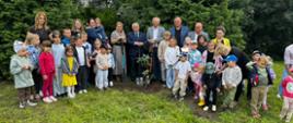 Grupa przedstawicieli władz i uczestników wydarzenia, w tym wicewojewoda Zbigniew Szczypiński, pozuje wspólnie z grupą przedszkolną przy posadzonym drzewie. Oficjele trzymają łopaty.