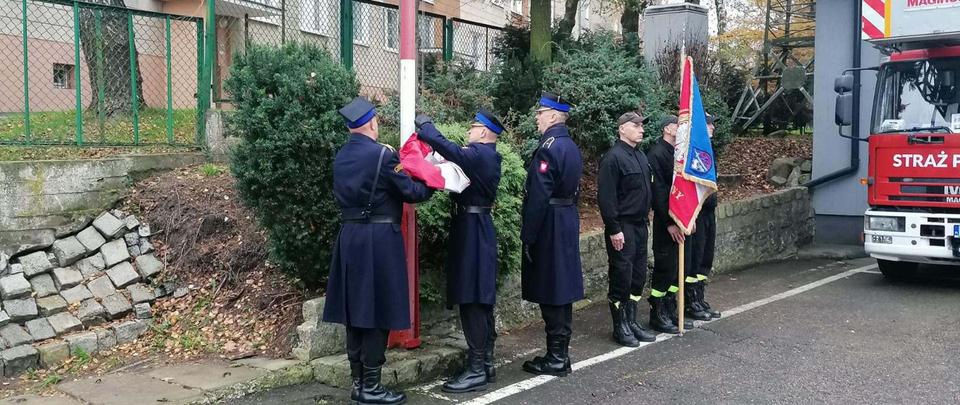 Plac wewnętrzny. Przed biało czerwonym masztem trzech strażaków w mundurach galowych podczepia flagę. Z prawej strony stoi trzech strażaków ze sztandarem. W prawym rogu samochód drabina.