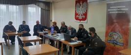 Zdjęcie przedstawia 7 mężczyzn i 1 kobietę siedzących za stołem. Na prawo za piątką osób na ścianie wisi godło Polski oraz rozstawiony jest rolap Komendy Powiatowej Państwowej straży Pożarnej, a za trójką osób siedzących na wprost jest okno z białymi firankami i szarymi zasłonami. Na stole przed nimi stoi laptop oraz dokumenty, które mężczyźni i kobieta czytają. Dwaj mężczyźni ubrani są w galowy strój osp, kolejni dwaj mężczyźni ubrani są w stroje dowódczo-sztabowe, reszta osób ubrana jest w stroje cywilne.