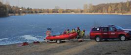 Na zdjęciu widać samochód strażacki z łodzią który został zadysponowany do zdarzenia. Strażacy przygotowują niezbędny sprzęt potrzebny do ewakuacji natomiast na tafli lodu po lewej stronie zdjęcia widać ratownika asekurowanego z brzegu na linie, który dotarł do poszkodowanego i pomaga mu w utrzymaniu się na powierzchni wody.