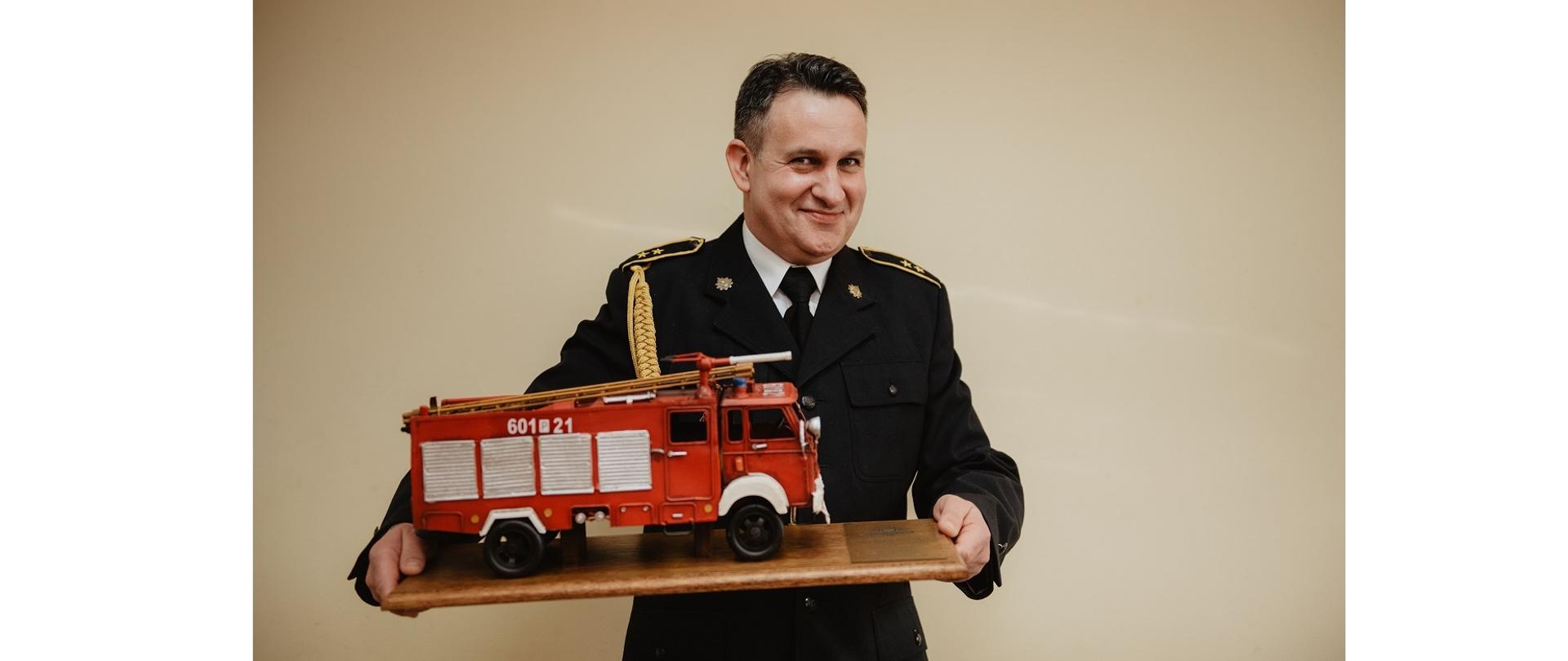 Zdjęcie przedstawia strażaka - st. asp. Marka Matuszaka w umundurowaniu wyjściowym trzymającego makietę czerwonego, strażackiego samochodu.