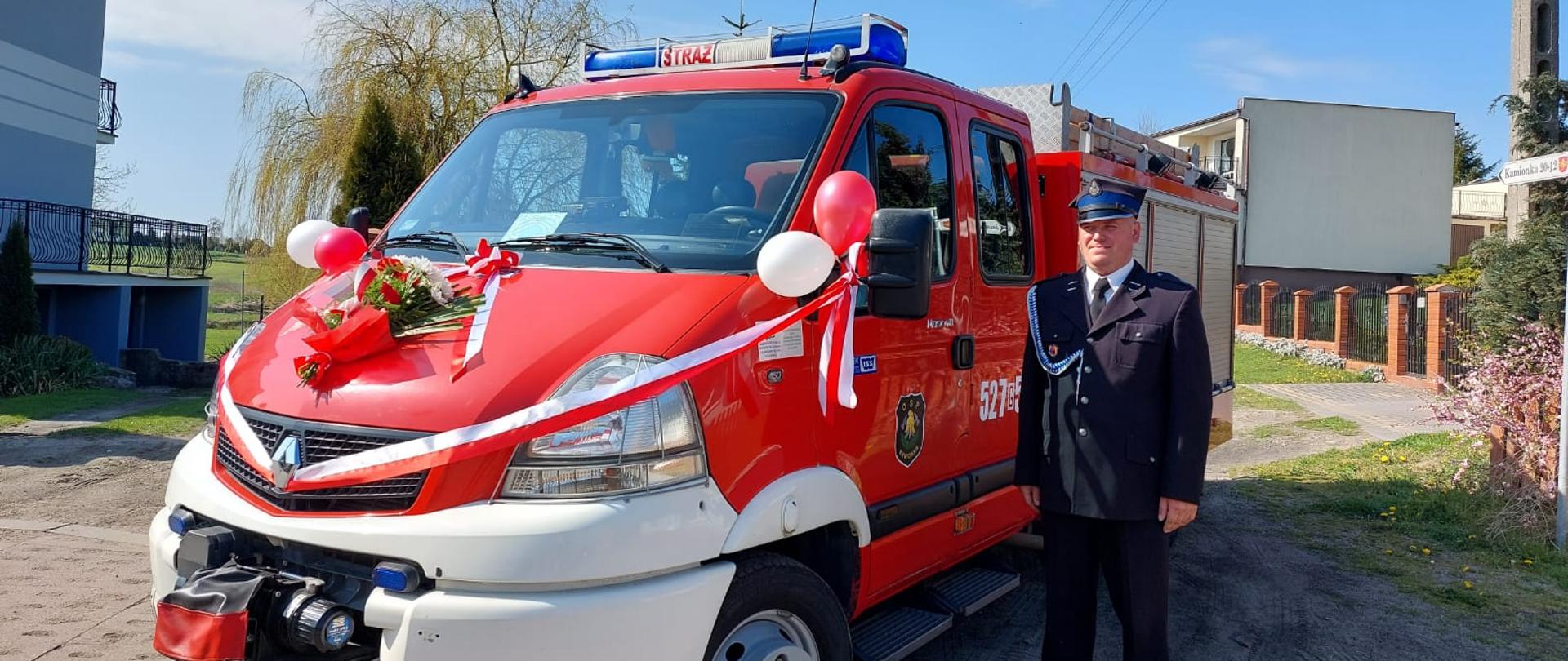 Zdjęcie przedstawia nowy pozyskany samochód pożarniczy, przepasany ozdobną szarfą z balonami. Z prawej strony stoi strażak w mundurze galowym