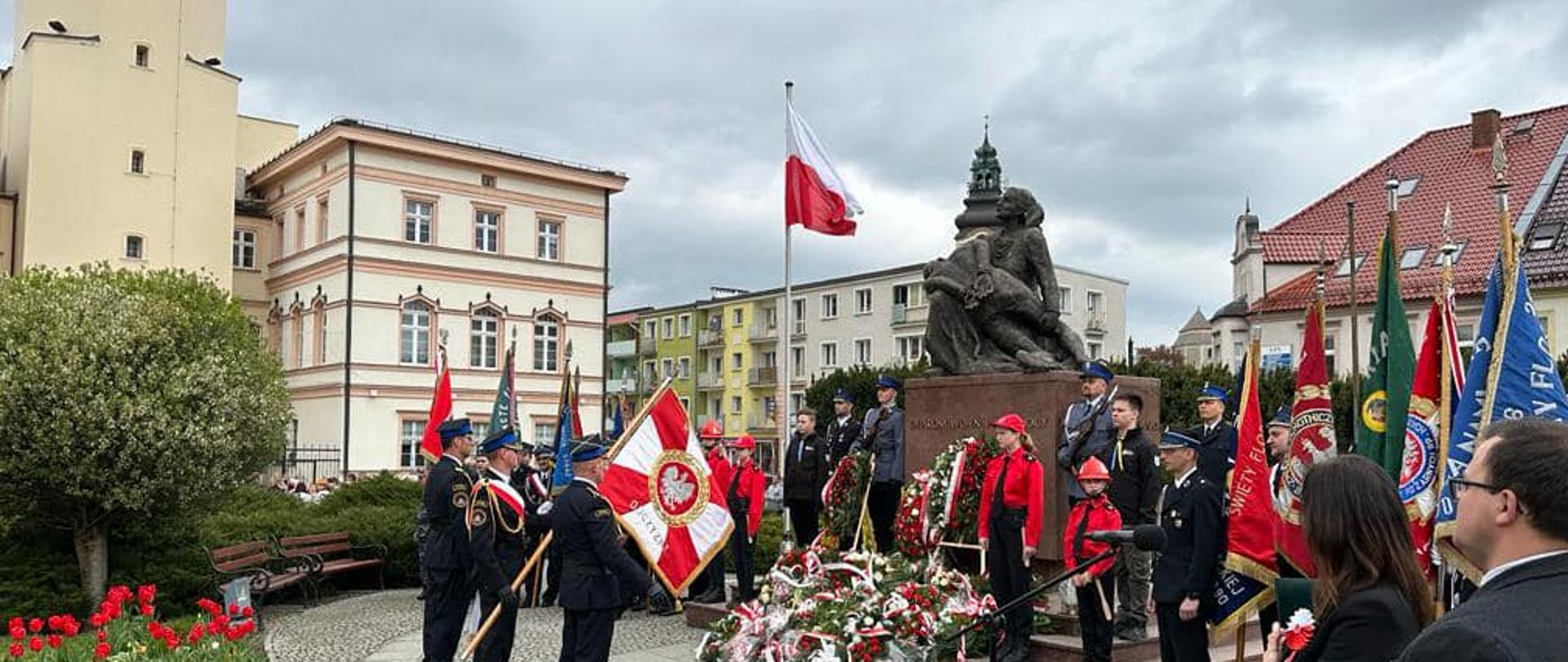 Na zdjęciu znajdują się poczty sztandarowe przy pomniku oraz warta honorowa, w tle widać budynki