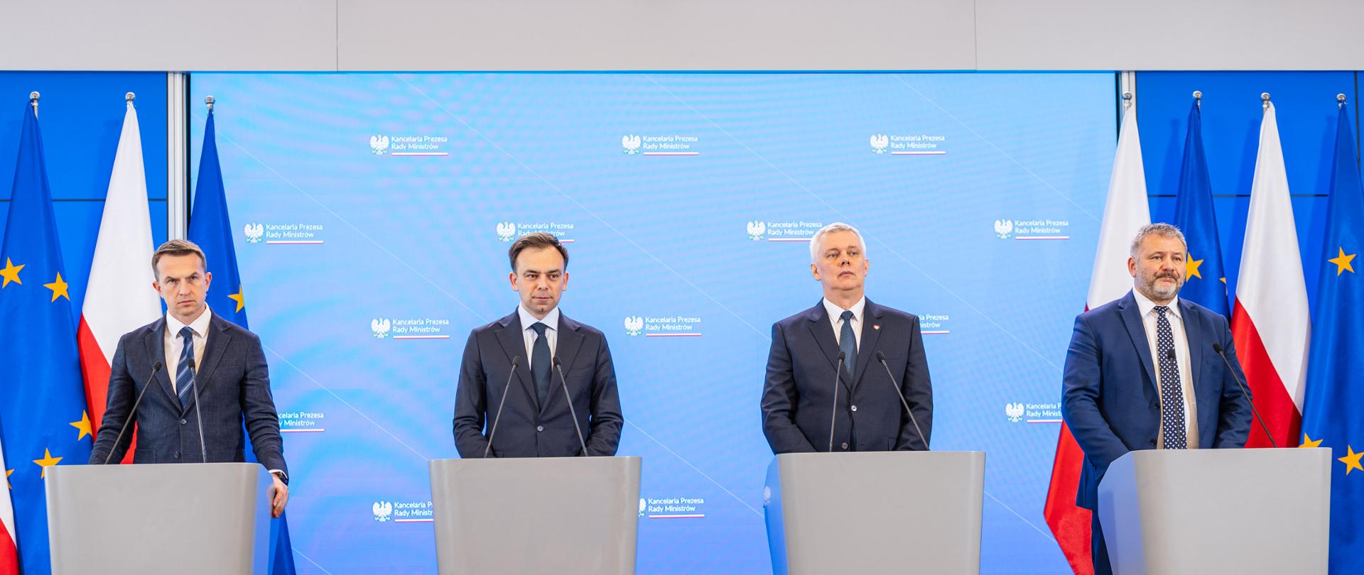 Zdjęcie przedstawia czterech ministrów podczas konferencji prasowej w KPRM - od prawej: minister Waldemar Żurek, minister Tomasz Siemoniak, minister Andrzej Domański, minister Adam Szłapka (rzecznik rządu). Ministrowie stoją za szarymi mównicami. W tle niebieska ścianka multimedialna z logo KPRM (napis Kancelaria Prezesa Rady Ministrów i orzeł). Po prawej i lewej stronie zdjęcia po 4 flagi - Polski (biało-czerwona) i UE (niebieska z żółtymi gwiazdkami).