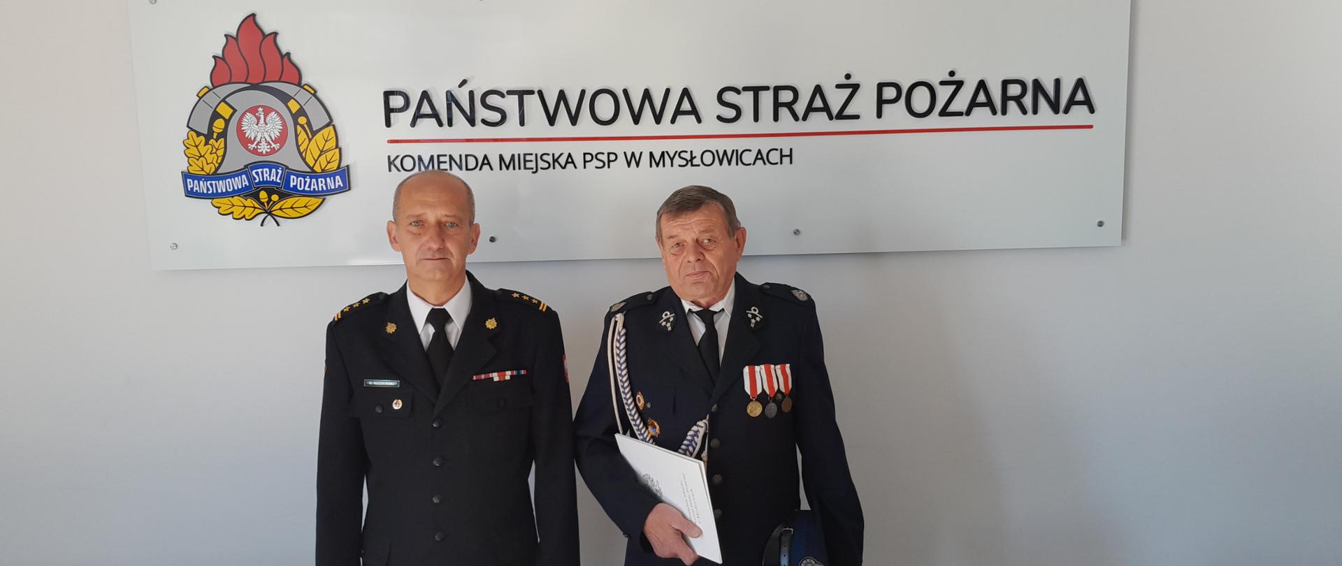 st. bryg. Olgierd Koźmiński wręcza decyzję w sprawie przyznania świadczenia ratowniczego druhowi OSP Janów z Mysłowic w tle logo PSP i napis Państwowa Straż Pożarna Komenda Miejska PSP w Mysłowicach