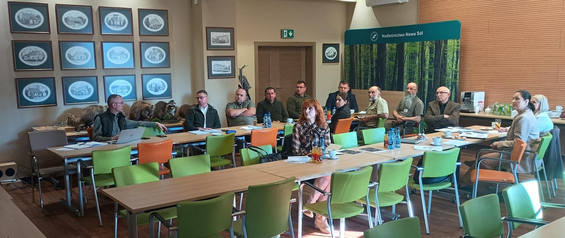 W dużej sali przy stołach siedzi kilkanaście osób.