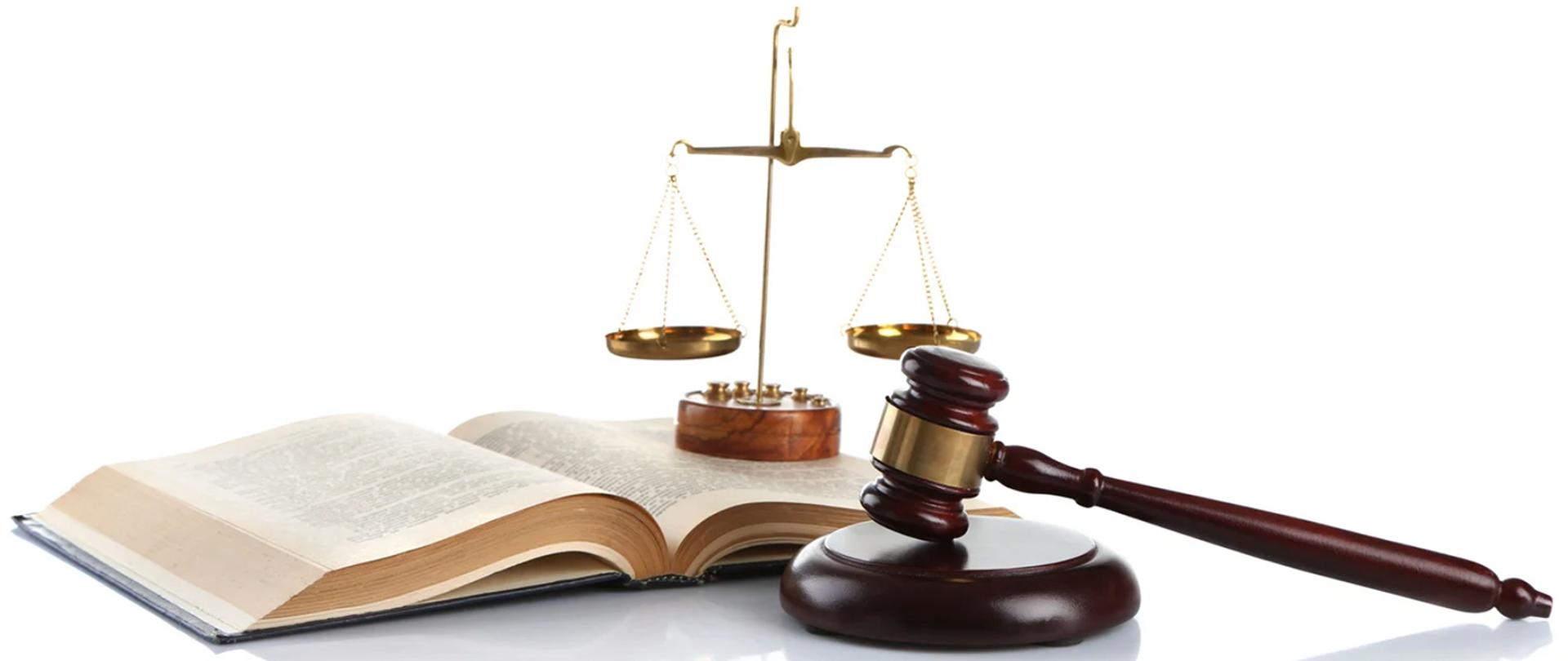 Na zdjęciu na białym blacie leży drewniany młotek sędziowski na podstawce, rozłożona w połowie księga prawa, na której postawiona została ozdobna złota waga - symbol sprawiedliwość.