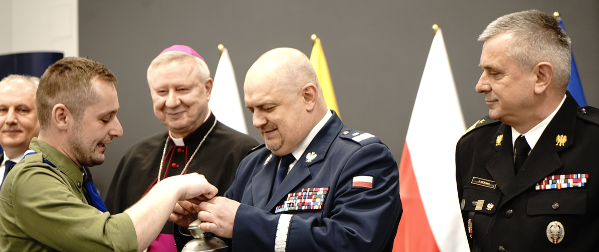 Harcerz przekazuje światełko betlejemskie komendantowi wojewódzkiemu Policji obok stoi pomorski komendant wojewódzki Państwowej Straży Pożarnej biskup i wojewoda pomorski za nimi stoi flaga Polski.