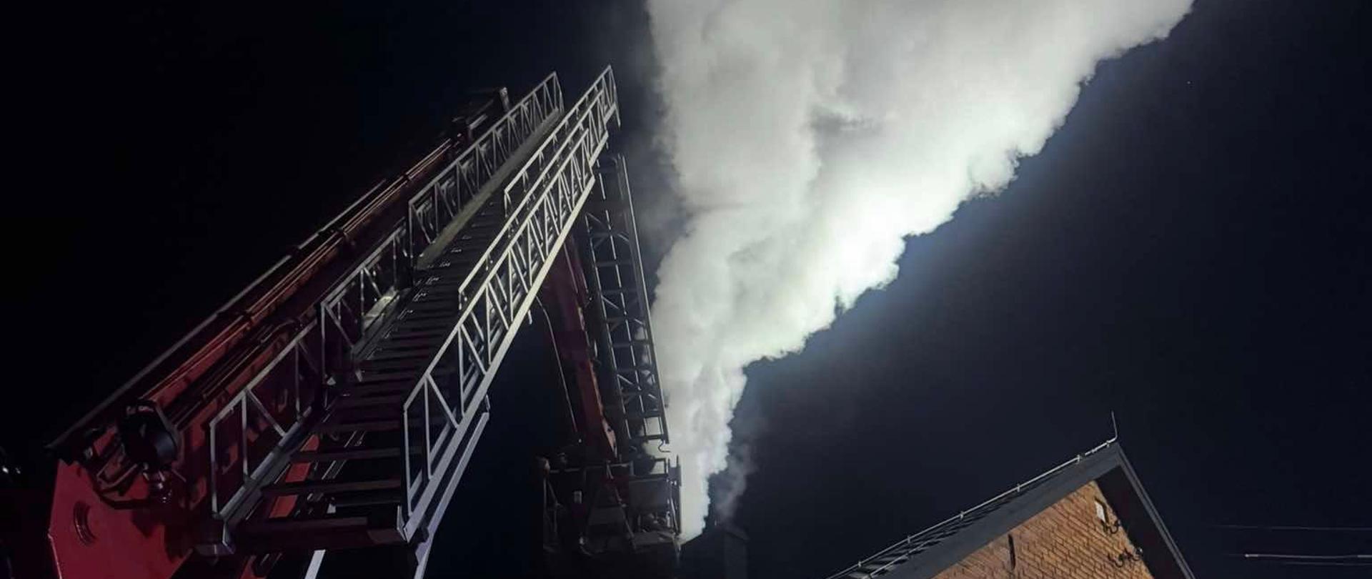 Budynek mieszkalny murowany z cegły. Z komina znajdującego na szczycie budynku unosi sie gęsty, jasny dym . Z lewej strony, na podwórzu stoi samochód strażacki z popdnośnikiem hydraulicznym. Obok niego są strażacy uczestniczący w akcji. 