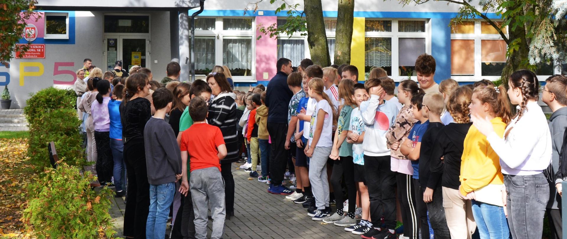 Grupa dzieci przed szkołą w punkcie zbiórki w trakcie ewakuacji.
