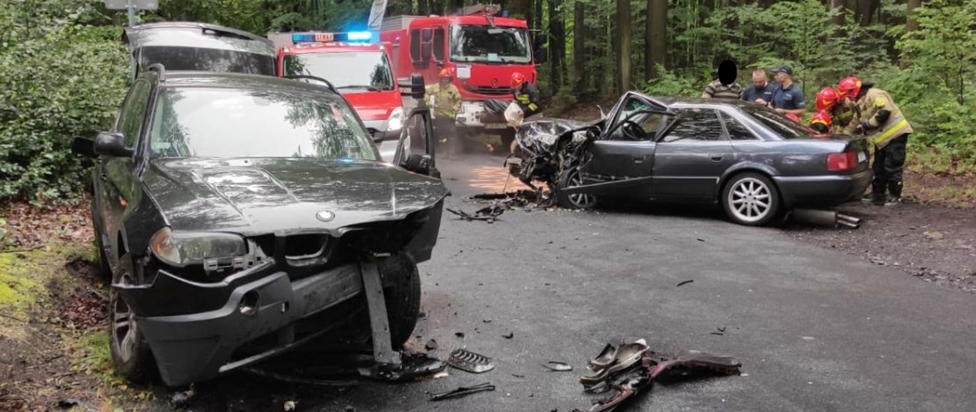 Na zdjęciu widać dwa samochody osobowe po zderzeniu czołowym. Jeden marki BMW a drugi Audi. Droga zablokowana w obu kierunkach. Na zdjęciu widać również dwa samochody straży pożarnej i strażaków podczas wykonywania czynności związanych z wypadkiem i udzielaniem pierwszej pomocy przedmedycznej.