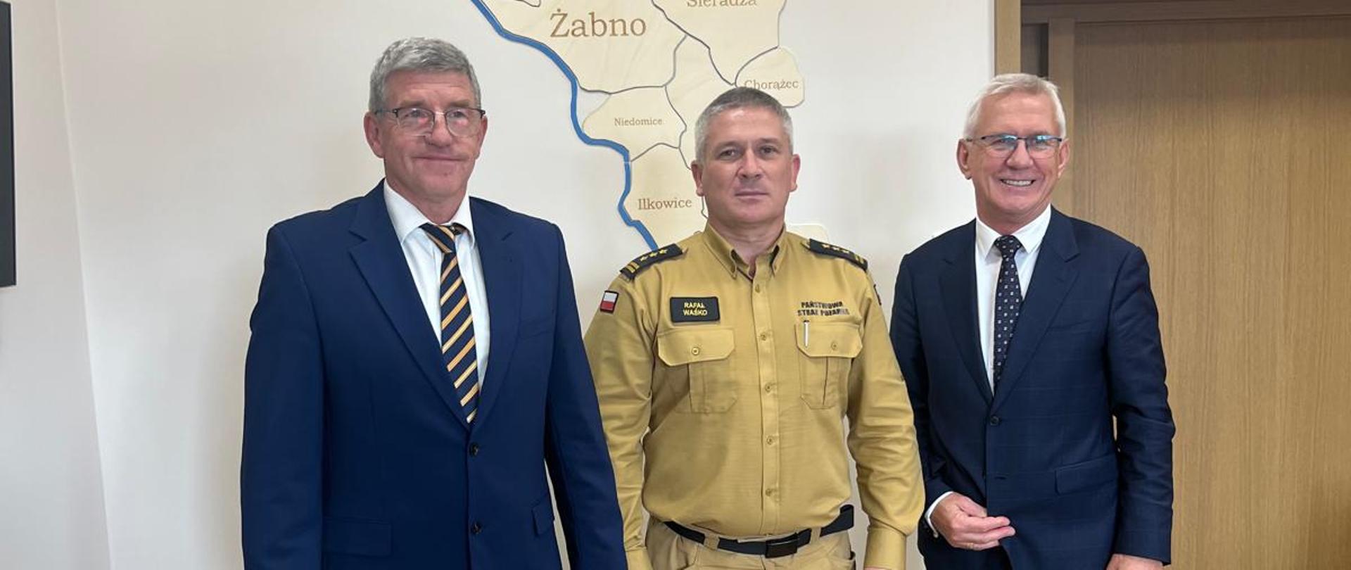 Trzy osoby stoją obok siebie, osoba w środku ubrana jest w piaskowy mundur strażacki, natomiast pozostałe osoby ubrane są w granatowe garnitury.