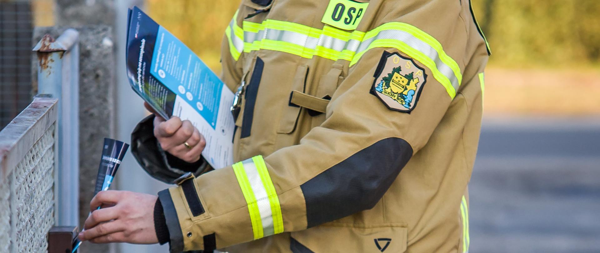 Zdjęcie przedstawia strażaka w ubraniu specjalnym, który w ręku trzyma ulotki i jedną z nich wrzuca do skrzynki na listy