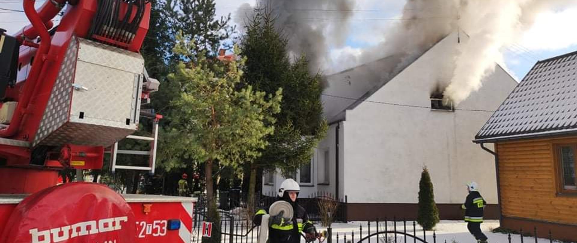 Na zdjęciu widać wydobywający się dym z poddasza oraz dachu w budynku mieszkalnym jednorodzinnym w Kunowie. Na pierwszym planie widać strażaka przy samochodzie strażackim, który z wężem w ręku przystępuje do jego rozwinięcia. W tle pożar budynku.