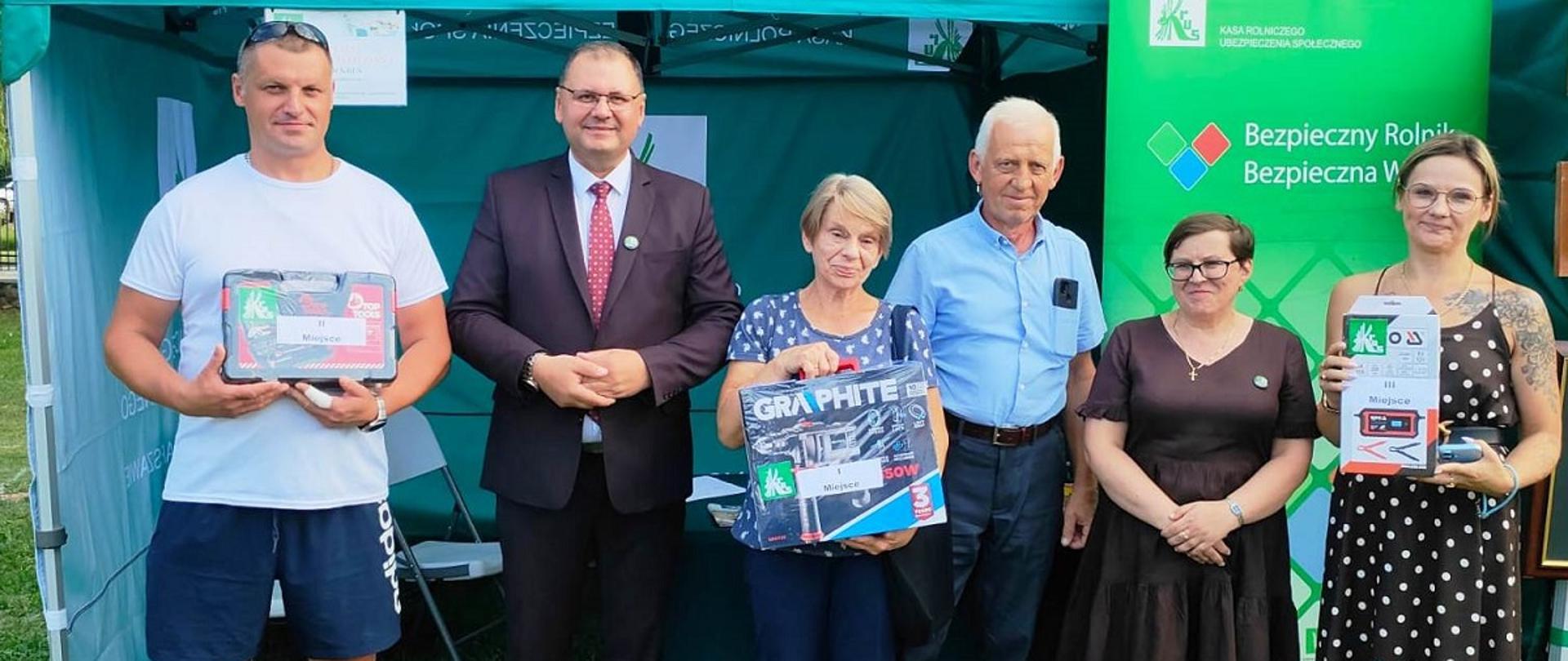 Grupa sześciu osób stoi przed zielonym namiotem z logo KRUS i napisem Kasa Rolniczego Ubezpieczenia Społecznego. Część z nich trzyma nagrody, na których są naklejki z logo KRUS. Po prawej stronie widoczny jest zielony baner logo KRUS i hasłem „Bezpieczny Rolnik, Bezpieczna Wieś”.