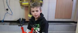 Remiza OSP. Jeden z uczestników eliminacji trzyma w rękach stojak hydrantowy. Góra urządzenia jest czerwona. Chłopiec ubrany jest w ciemną bluzę z krótkim rękawem, spod której wystają białe rękawy innej bluzy. W tle - wieszak, na którym zawieszone są ubrania specjalne w kolorze piaskowym.