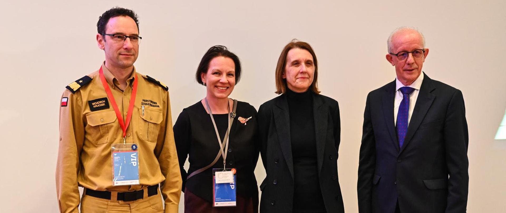 Cztery osoby stoją na tle jasnej ściany, pozując do wspólnego zdjęcia. Po lewej stronie znajduje się mężczyzna w mundurze Państwowej Straży Pożarnej z identyfikatorem VIP i napisem "Wojciech Kruczek". Obok niego stoją dwie kobiety w eleganckich strojach oraz starszy mężczyzna w garniturze i okularach. Wszyscy się uśmiechają.