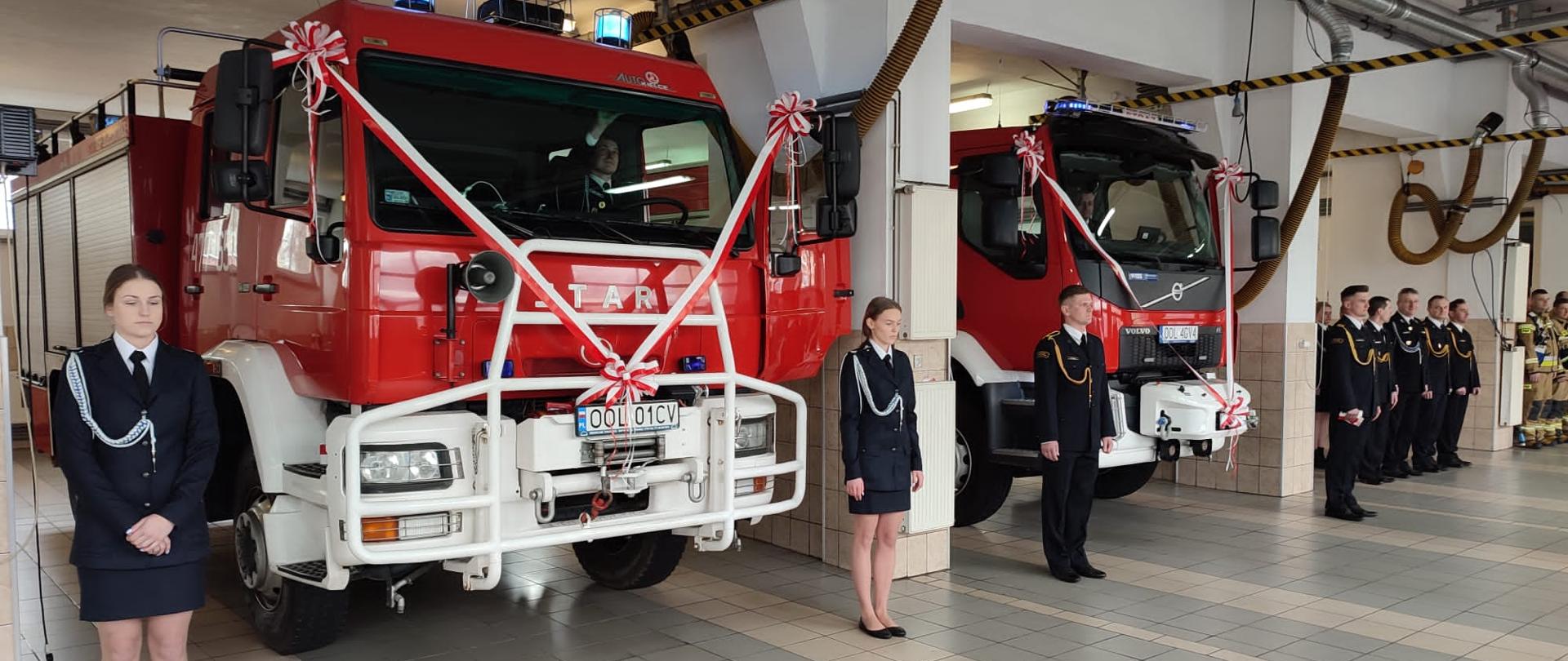 W garażu stoją dwa samochody pożarnicze koloru czerwonego. Obok samochodów stoją strażacy w mundurach wyjściowych.