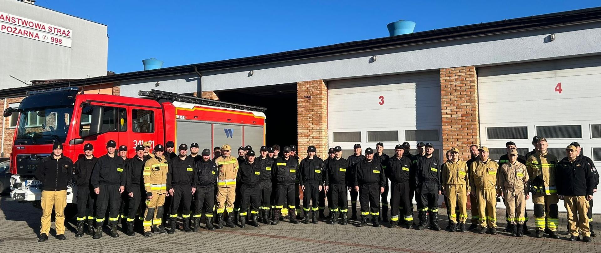 Na zdjęciu widać grupę strażaków stojących przed garażem straży pożarnej. W tle widoczne są garaże z numerami 3 i 4 oraz czerwony wóz strażacki. Uczestnicy ubrani są w mundury koszarowe i bojowe z odblaskowymi elementami oraz naszywkami „STRAŻ”. Część z nich ma na sobie czarne mundury, inni w kolorze piaskowym. Nad budynkiem widać napis „PAŃSTWOWA STRAŻ POŻARNA”. Zdjęcie wykonano w słoneczny dzień. Grupa podczas wspólnej fotografii pamiątkowej po zakończeniu szkolenia podstawowego strażaków OSP.